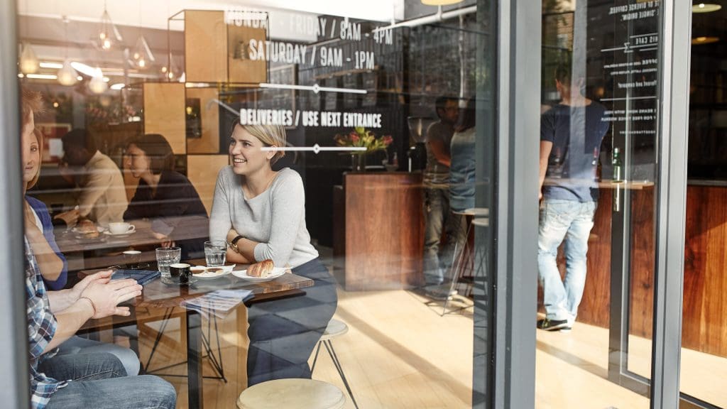 A woman sits at a table in a cozy café, smiling and conversing with a man across from her. Other patrons can be seen inside the café. The setting has a modern, inviting atmosphere with warm lighting. A menu board and glass windows highlight the café's interior.