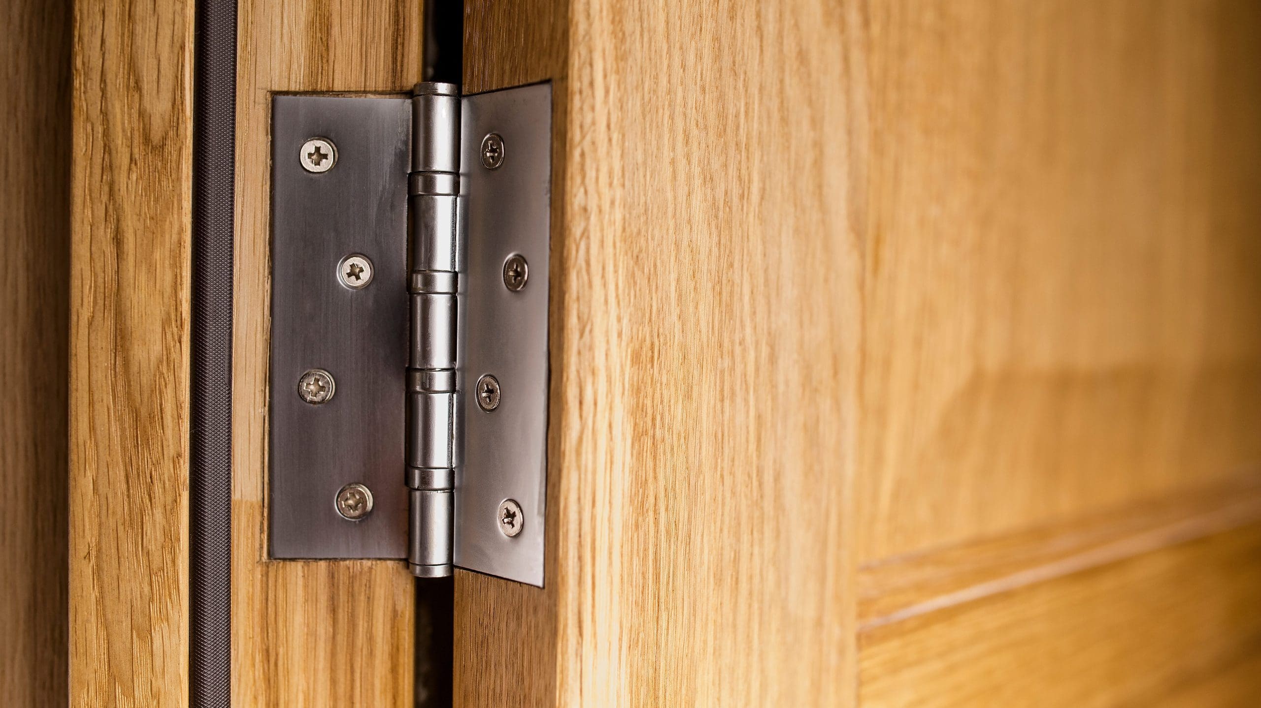 A close-up of a stainless steel hinge attached to a wooden door and door frame. The hinge has several screws securing it tightly, and the wood has a light, polished finish. The door is partially open, creating a narrow gap between the door and the frame.