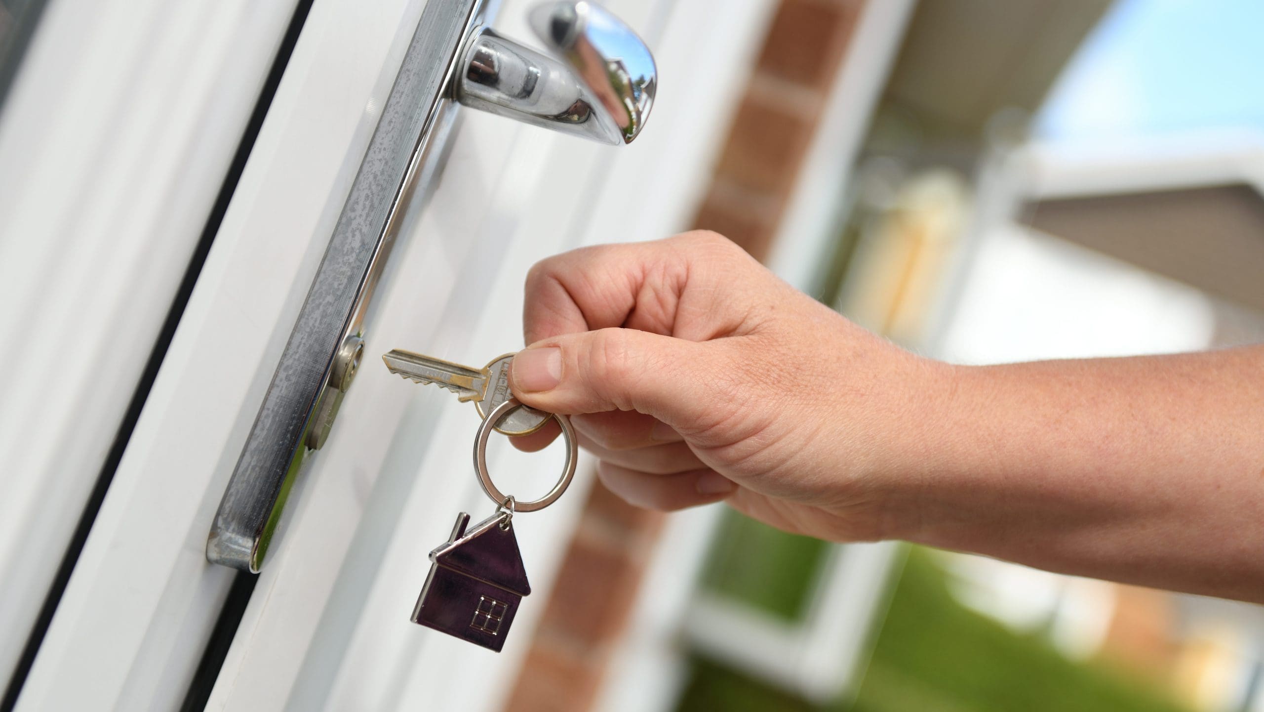 A person is inserting a key into a door lock with their right hand. The keychain features a small house-shaped pendant. The door appears to be white with a metal handle. The background is slightly blurred, showing part of a building and green foliage.