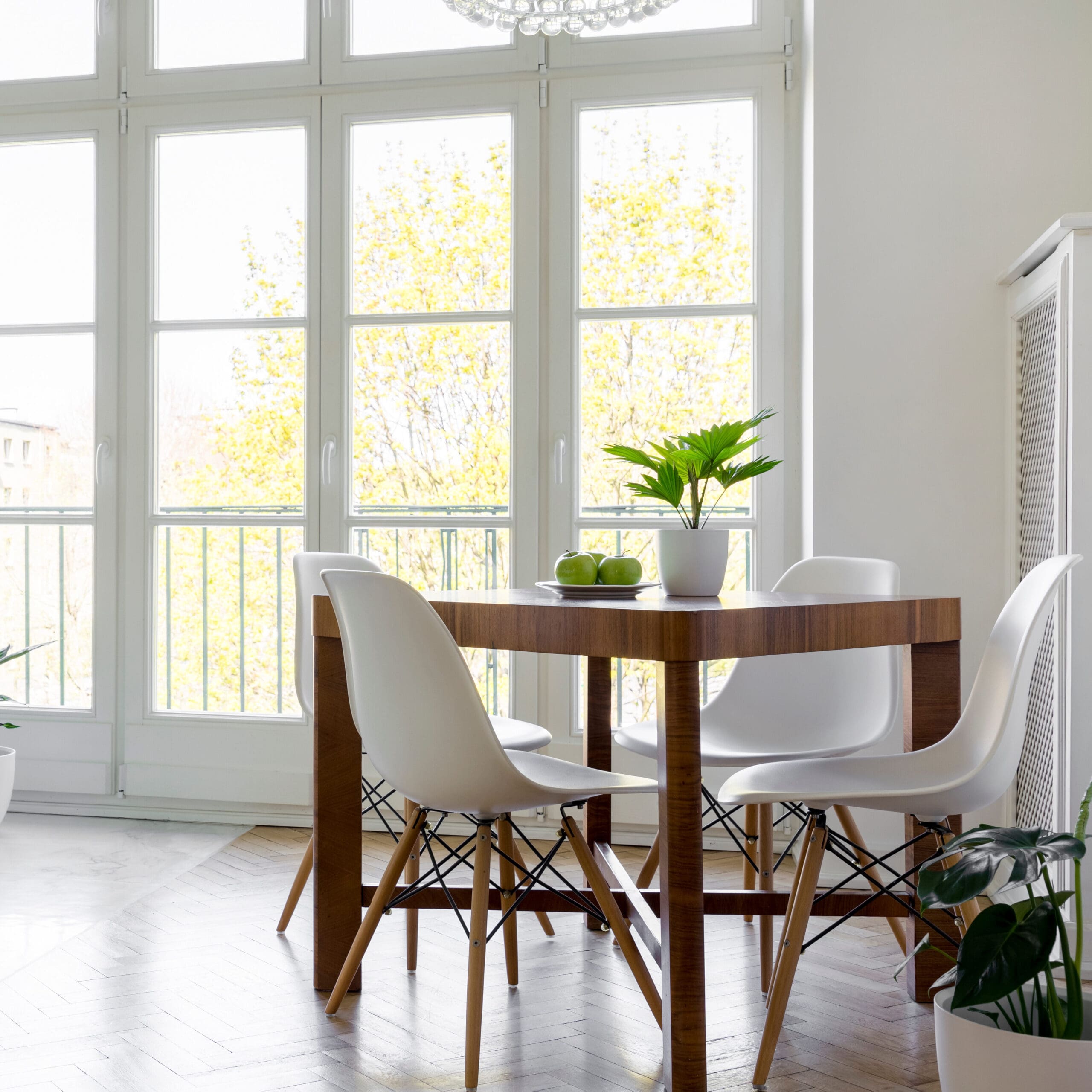 A modern dining area with a wooden table and four white chairs. A potted green plant and a bowl of apples sit on the table. Floor-to-ceiling windows in the background offer a view of trees with yellow leaves, allowing natural light to fill the room.