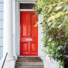 A bright red door with a mail slot is framed by white columns and set in a light blue exterior wall. A leafy green bush grows to the right of the door, partially obscuring the view. Steps lead up to the door, and a black iron fence runs along the garden path.