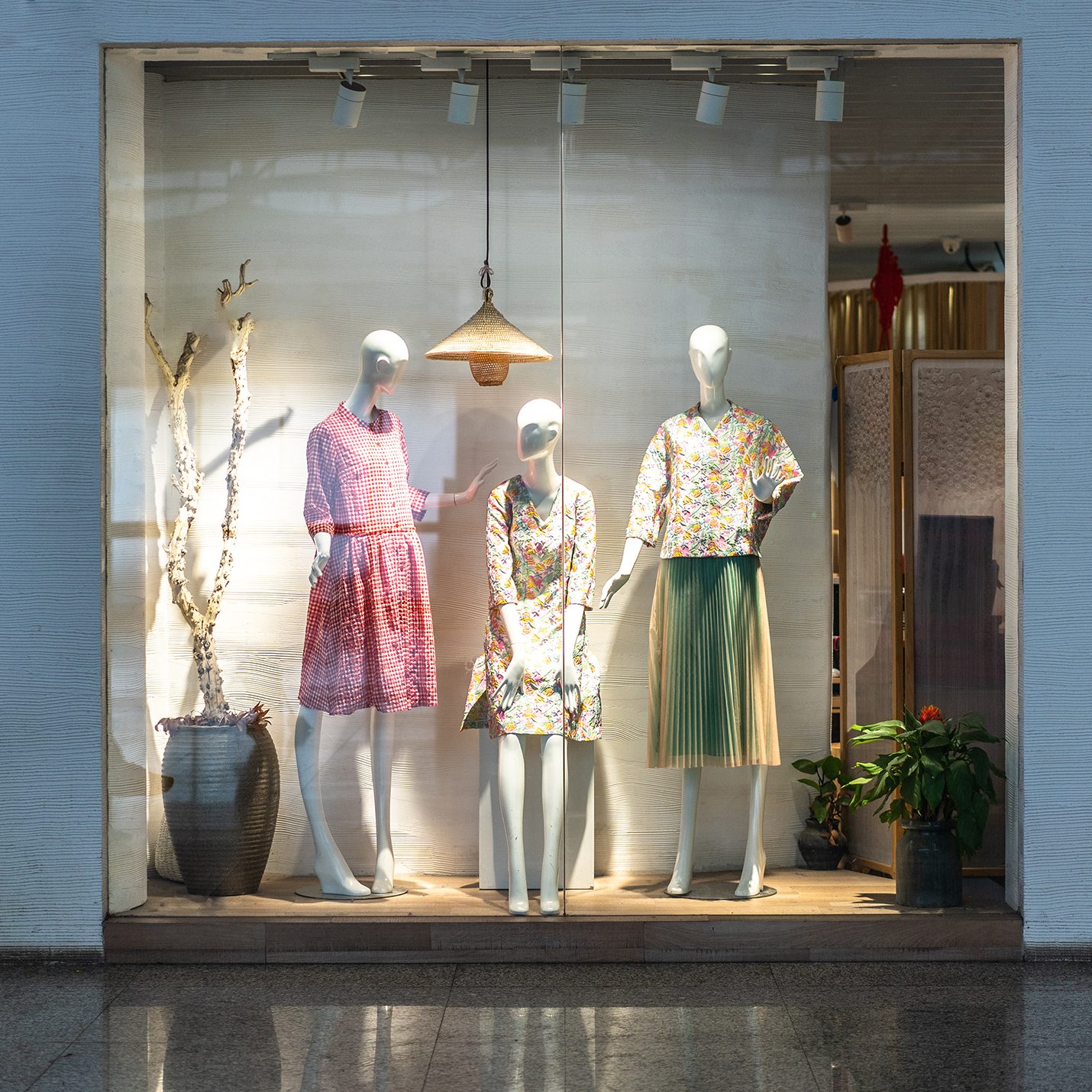 A store window display featuring three mannequins wearing colorful spring outfits is illuminated by a warm overhead light. The backdrop includes a large potted plant to the left and additional decorative elements, giving the display a fresh, natural feel.