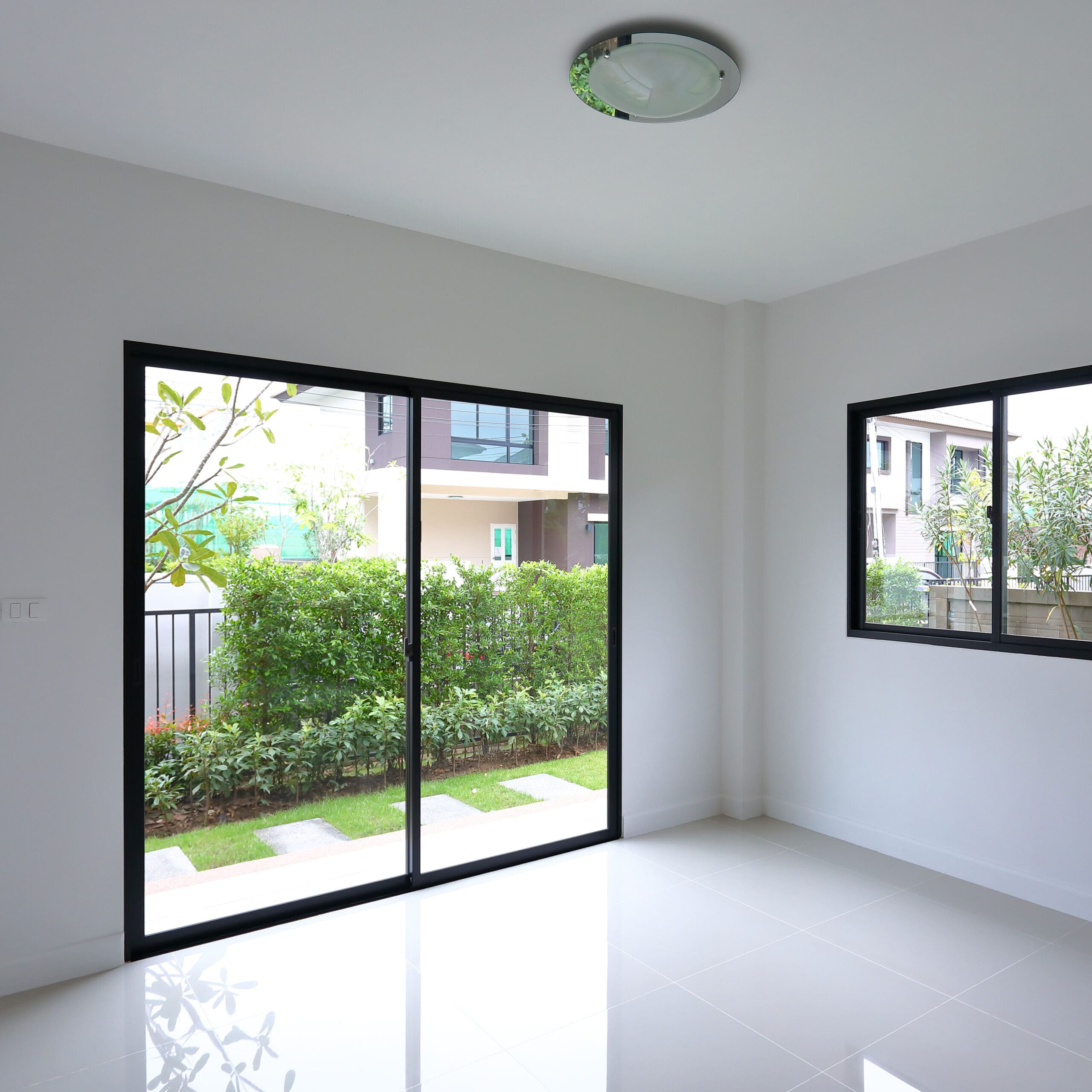 A bright, empty room with white walls and shiny tiled flooring. The room features a large sliding glass door and a window with black frames, both offering a view of a neatly trimmed garden and other modern houses outside. A round light fixture is on the ceiling.