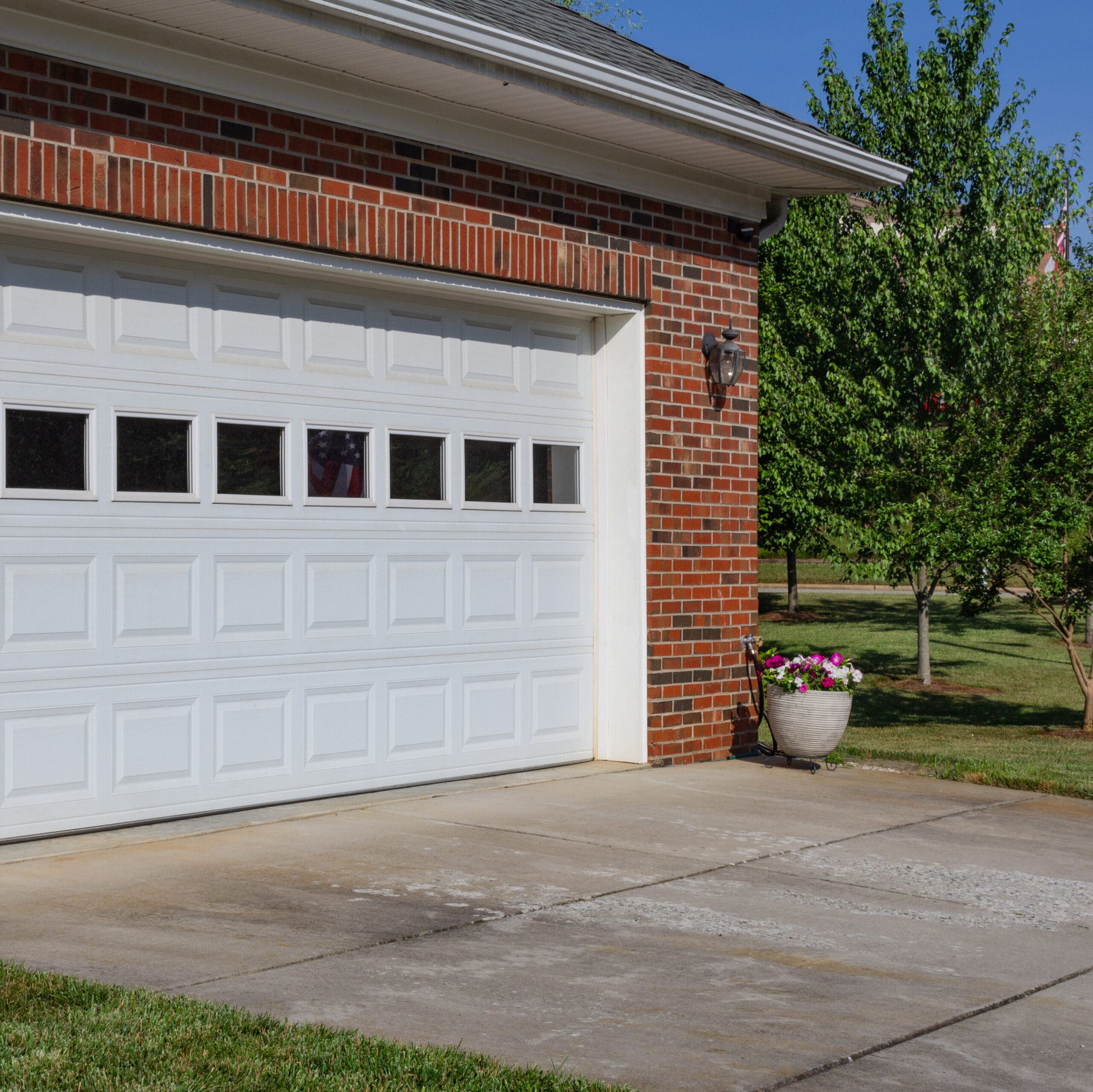 A brick garage with a white paneled door featuring five small windows in the upper row. A potted plant with pink and white flowers sits to the right of the door, next to a concrete driveway. Lush green trees are visible in the background under a clear blue sky.