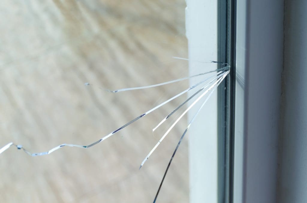 A close-up image of a cracked glass window. Several shards of glass are protruding from the point of impact, which is on the right side of the window. The background is blurred and light in color.