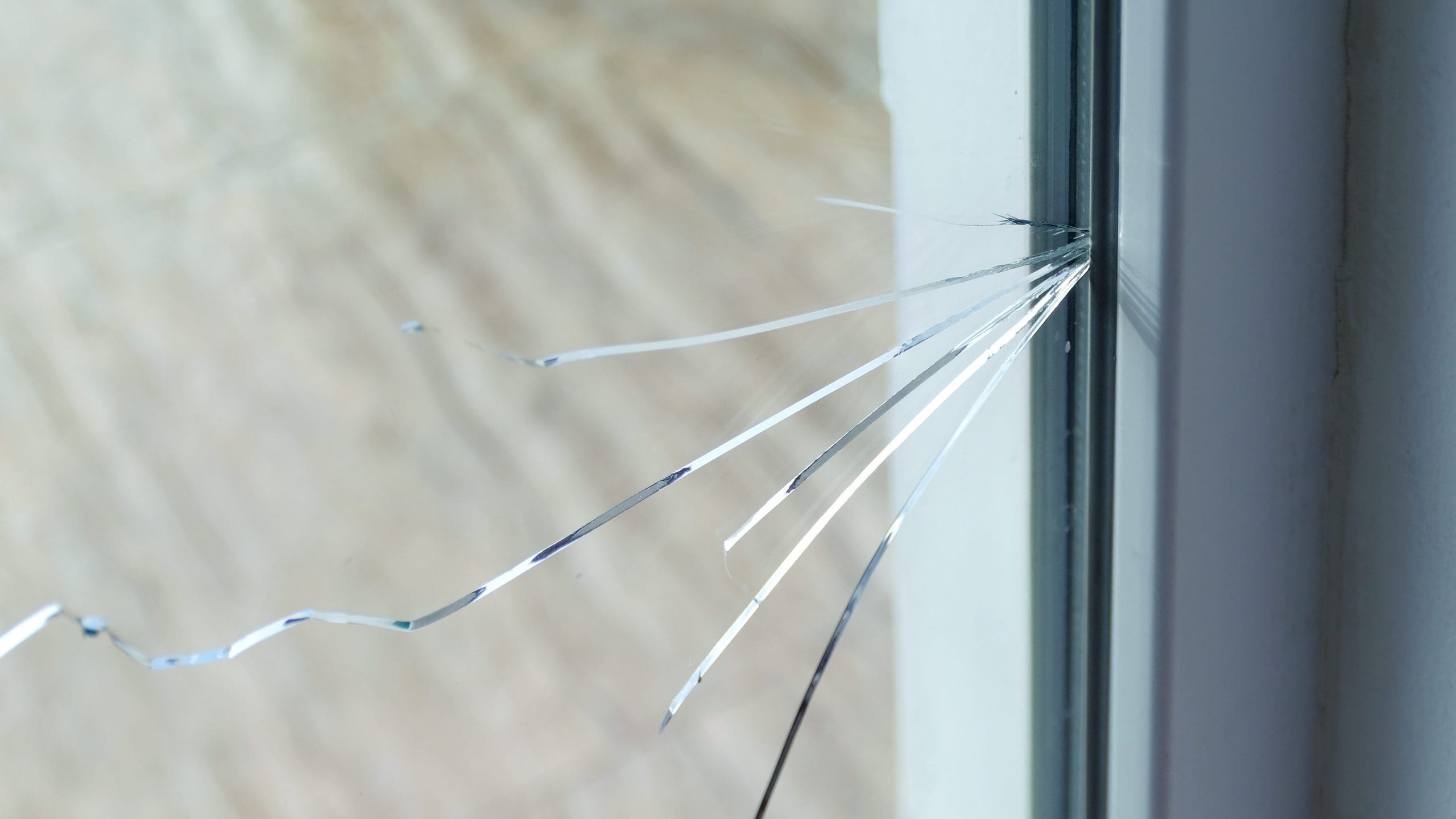 A close-up of a cracked glass window pane. Multiple thin cracks radiate outwards from a central point on the right side of the glass. The background is blurred, highlighting the damage on the window.