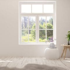 A bright, minimalist room with a large window showcasing a lush green outdoor view. The room features light wood flooring, a white pouf, and a small wooden table holding a vase with green and white flowers. Soft natural light fills the space.