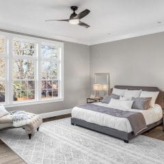 A serene bedroom with a large window, light gray walls, and hardwood floors. The room features a gray upholstered bed with white and gray bedding, two mirrored nightstands with table lamps, an area rug with geometric patterns, and a chaise lounge near the window.