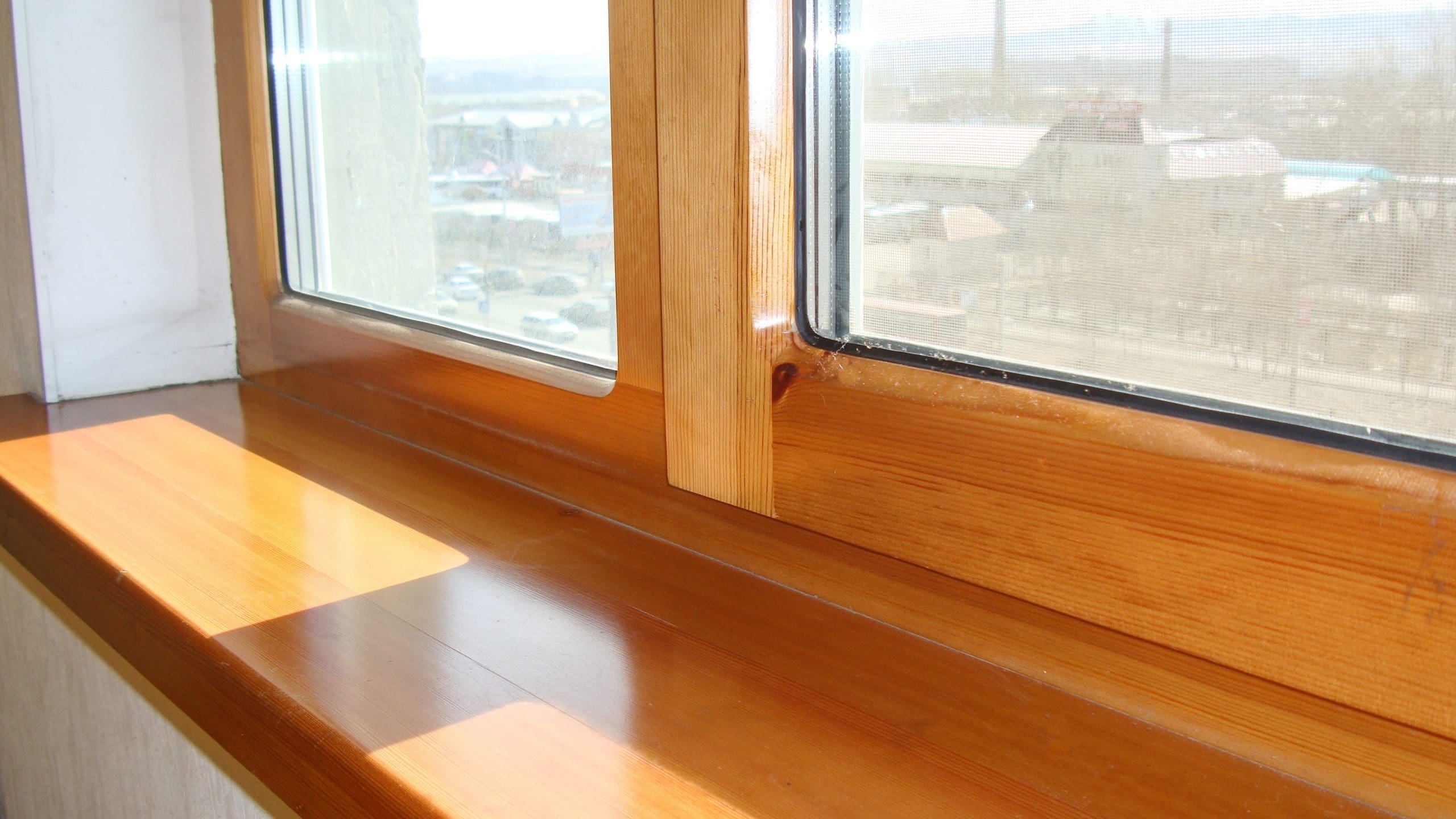 A close-up view of a wooden window sill and frame, with sunlight streaming through the glass. Outside the window, an urban landscape with buildings and streets can be faintly seen in the background. The scene appears bright and sunny.