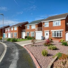 A row of two-story brick houses with neatly manicured lawns and shrubs on a suburban street under a clear blue sky. The homes have white trim and are situated along a curved road with a grassy verge and lampposts.