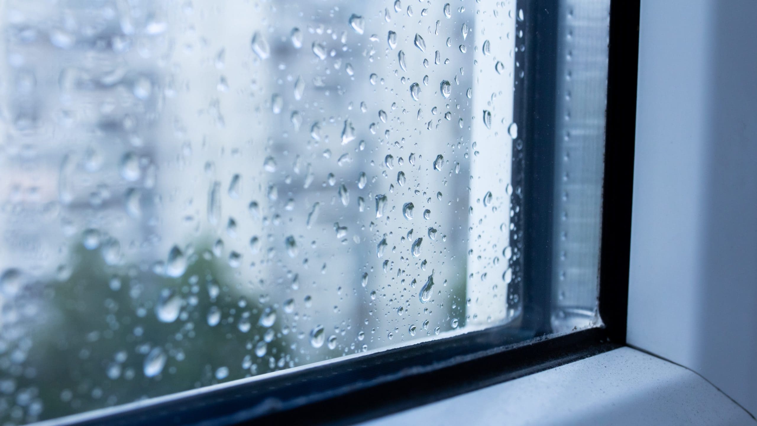 Close-up of raindrops on a windowpane, with water droplets indicating recent rainfall. The background appears blurred, with indistinct shapes suggesting buildings and greenery outside. The scene evokes a rainy day atmosphere viewed from inside.