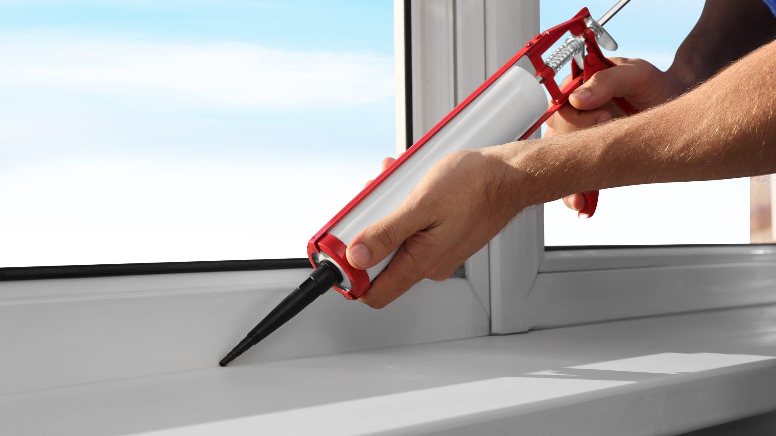 A person uses a red caulking gun to apply sealant around the edge of a white window frame. The background shows a clear, blue sky. The scene appears to be indoors, indicating home improvement or maintenance work.