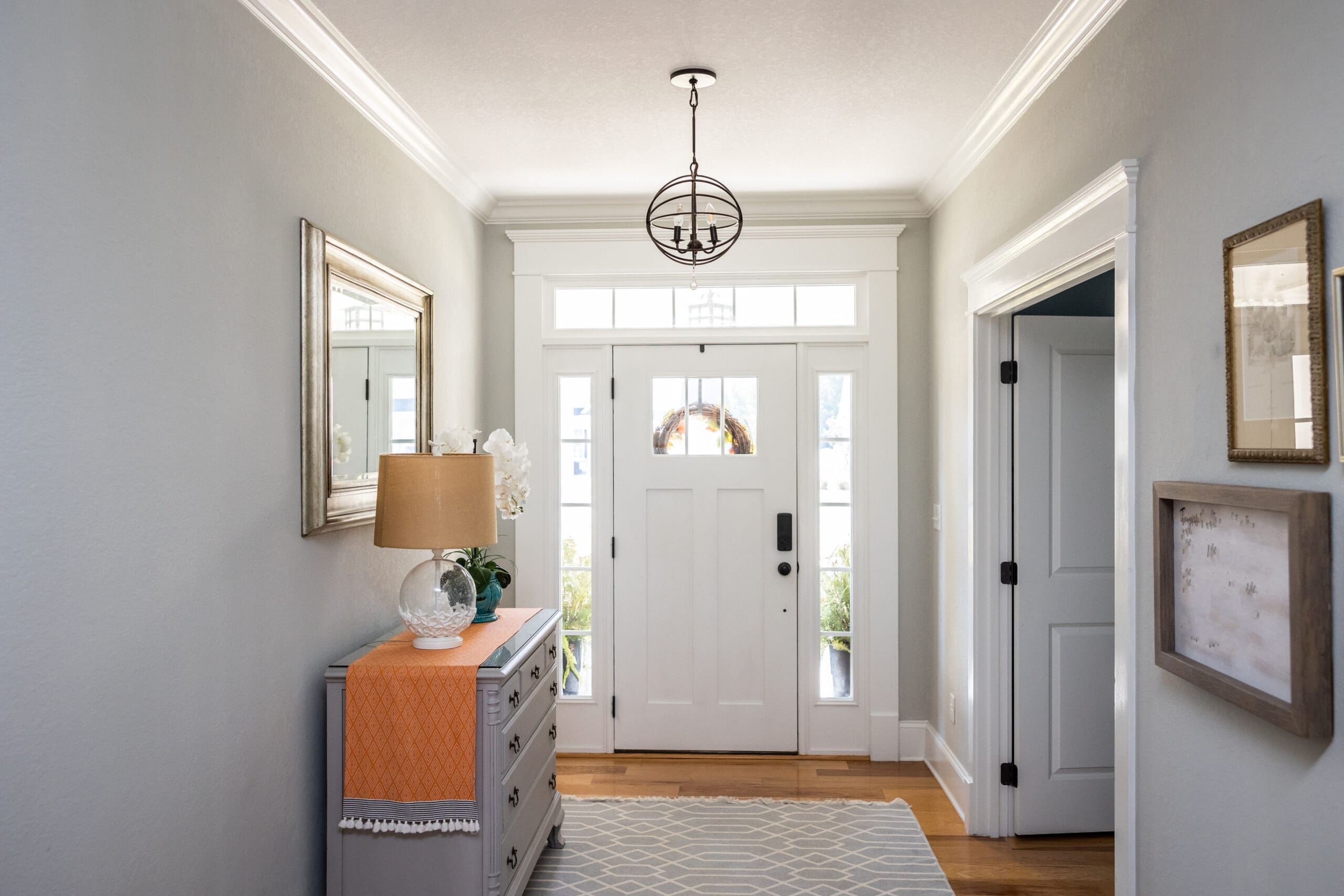 A bright and welcoming hallway with light gray walls, a white front door with glass panels, and hardwood flooring. The space features a gray console table with an orange runner, a lamp, a framed mirror above it, and a window allowing natural light.