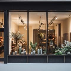 A street view of a modern florist shop with large glass windows showcasing various plants and floral arrangements. The interior is brightly lit, and several vases and potted plants are displayed on wooden shelves and tables inside.