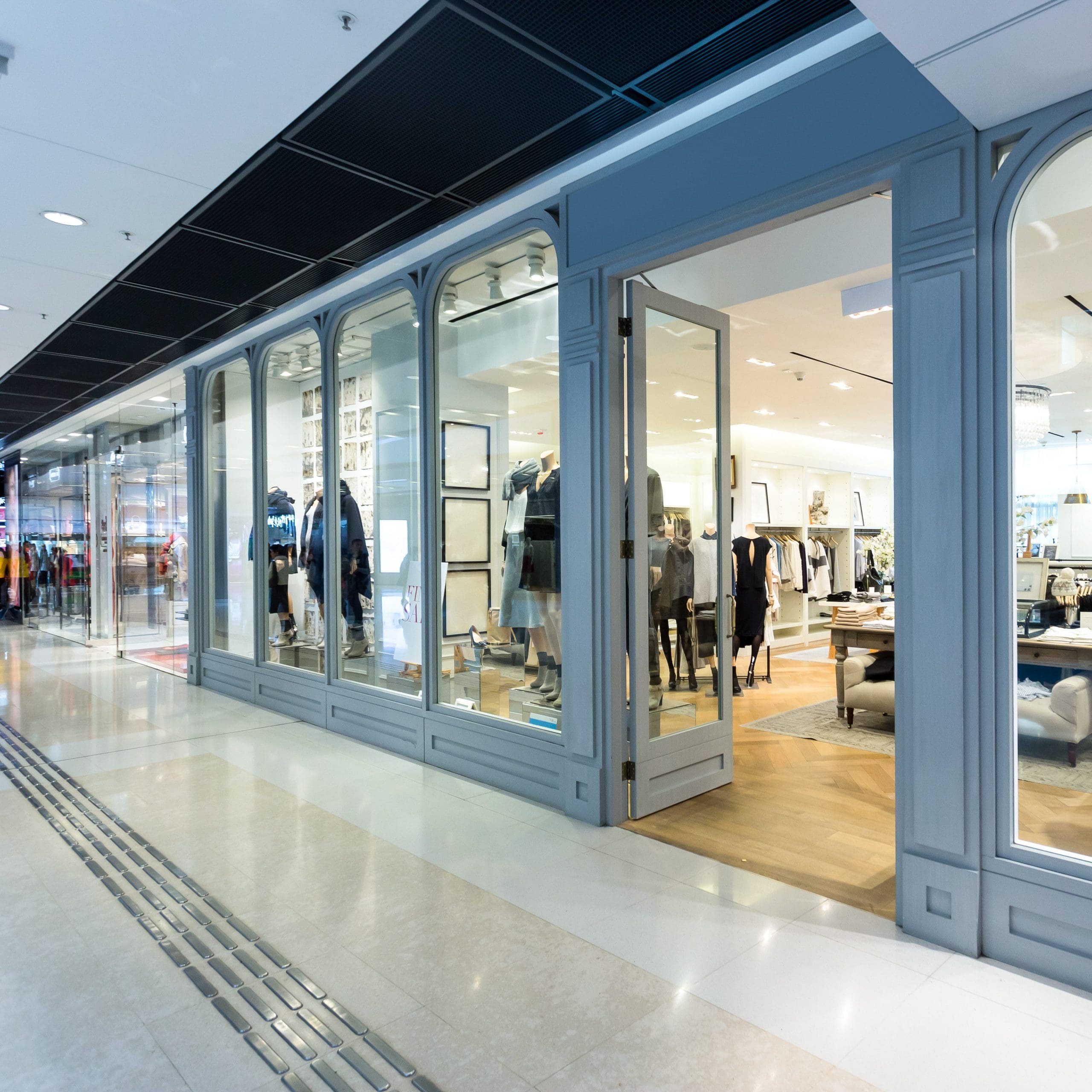 A modern clothing store in a shopping mall with large glass windows showcasing mannequins dressed in fashionable outfits. The store has a sleek, grey facade and well-lit interiors with clothing racks and shelves. The mall's corridor is visible in the foreground.