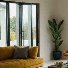 A modern living room with a large sliding glass door, a mustard-yellow couch with a gray pillow, and a tall green houseplant in a black pot. A wooden coffee table with a glass surface holds a tray with lemons and some books. Light streams in from outside.