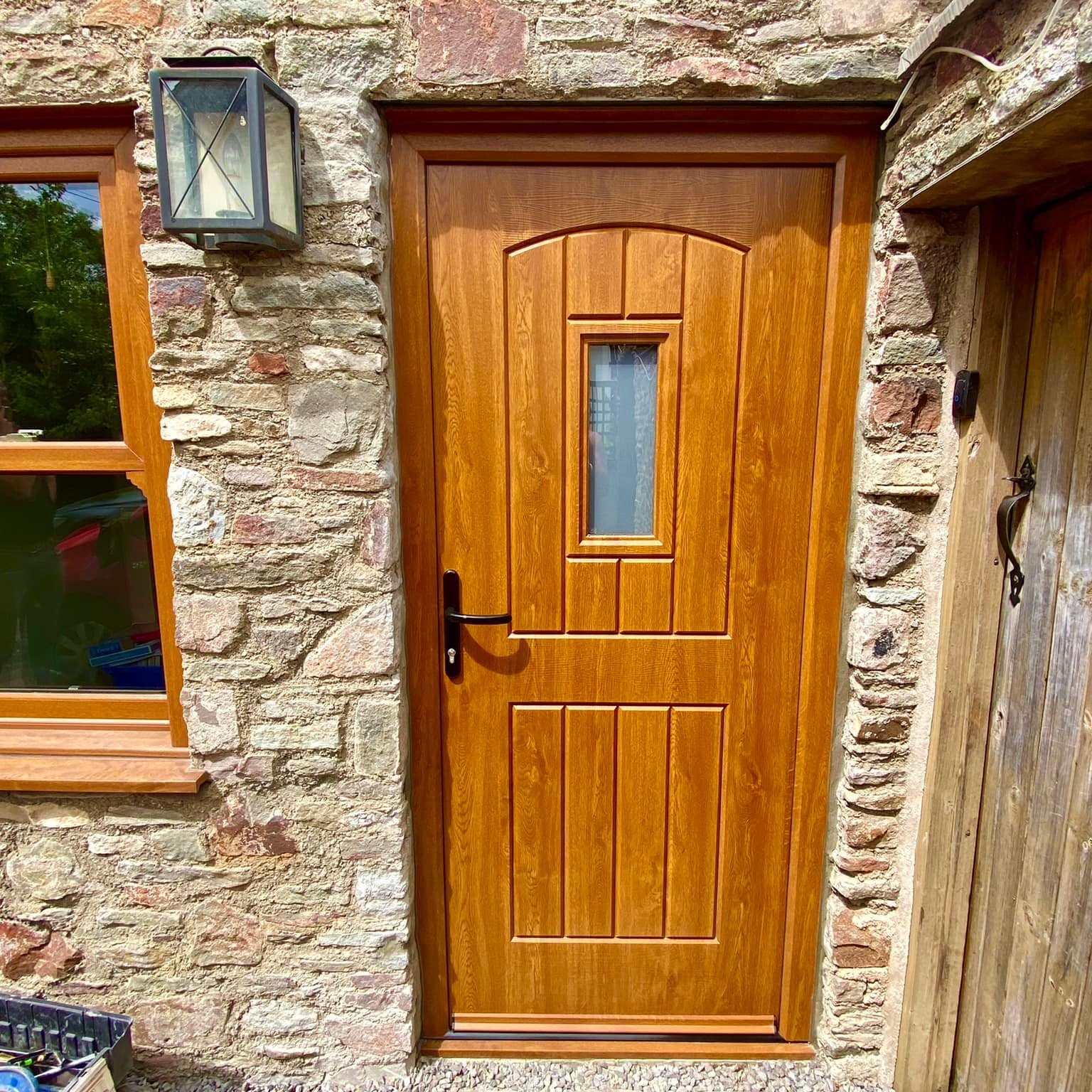 A wooden door with a small, rectangular, glass windowpane at the center is set in a rustic stone wall. To the left of the door, there is a small window with a wooden frame and a lantern-style light fixture above. A wooden structure is adjacent on the right.