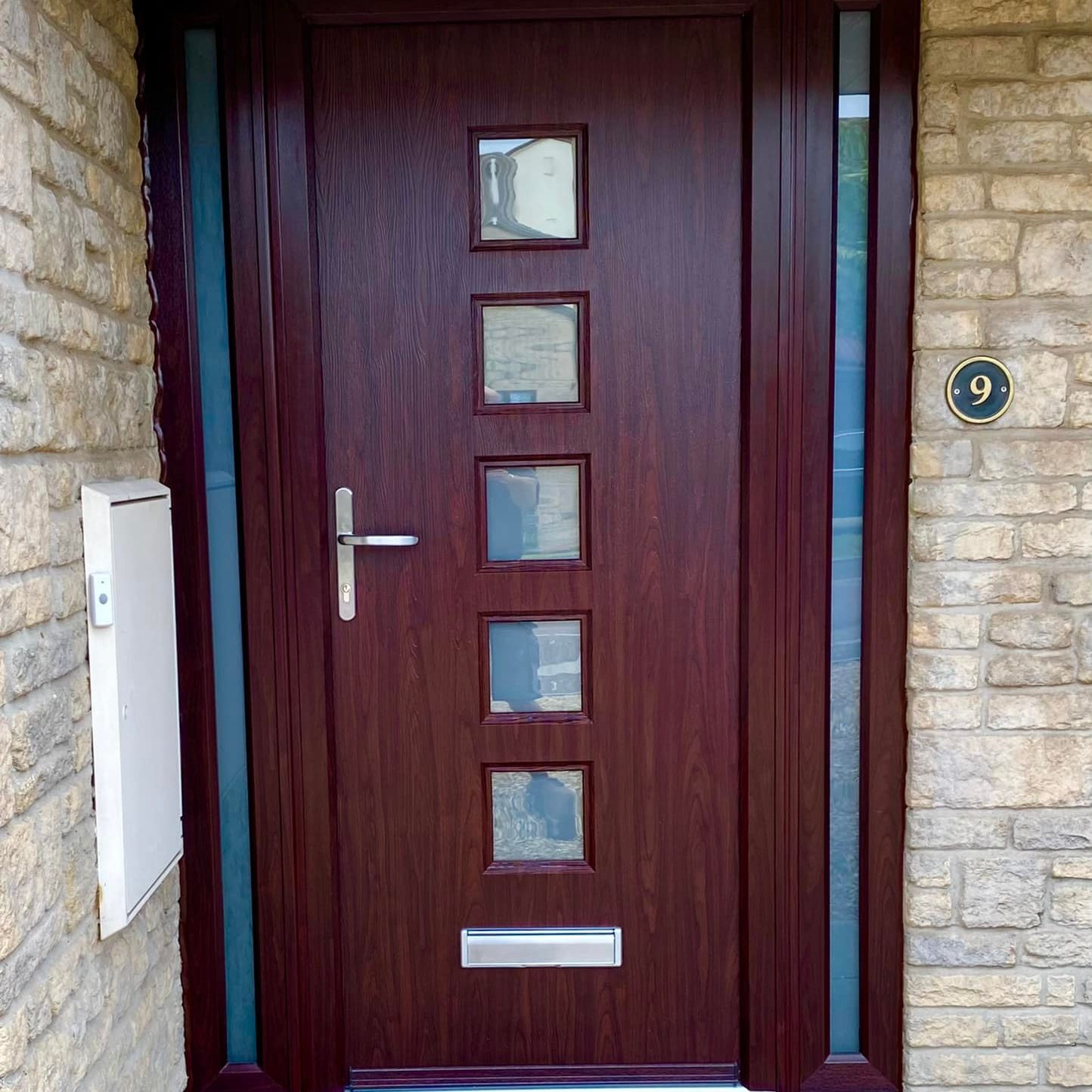 A dark red wooden door with four square glass panels arranged vertically in the center, framed by stone walls. The house number 9 is mounted to the wall on the right. A small mail slot is at the bottom of the door, and a welcome mat lies in front of it.