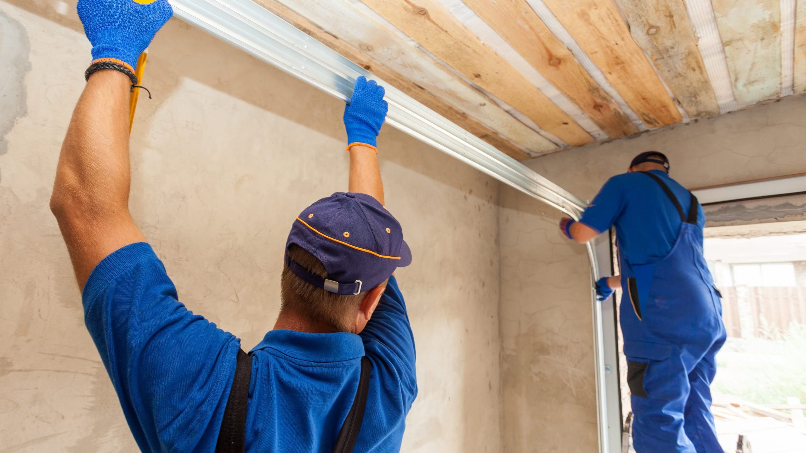 Two construction workers in blue uniforms and blue gloves are installing a metal frame. One worker is adjusting the frame at the top, while the other is securing it on the side. The room has bare, unfinished walls and a wooden ceiling.