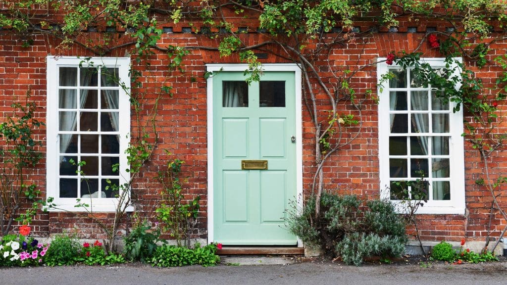 A charming brick house facade with a light green door, flanked by two white-paned windows. Framing the door and windows are climbing plants with green leaves, and there is a small garden bed with various flowers and shrubs in front of the house.