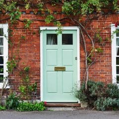 A charming brick house facade with a light green door, flanked by two white-paned windows. Framing the door and windows are climbing plants with green leaves, and there is a small garden bed with various flowers and shrubs in front of the house.