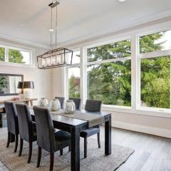 Modern dining room with large windows offering a view of lush greenery. Room features a black dining table surrounded by six gray upholstered chairs, a woven rug, and a wooden side table with a lamp and mirror. Ceiling has a rectangular chandelier.