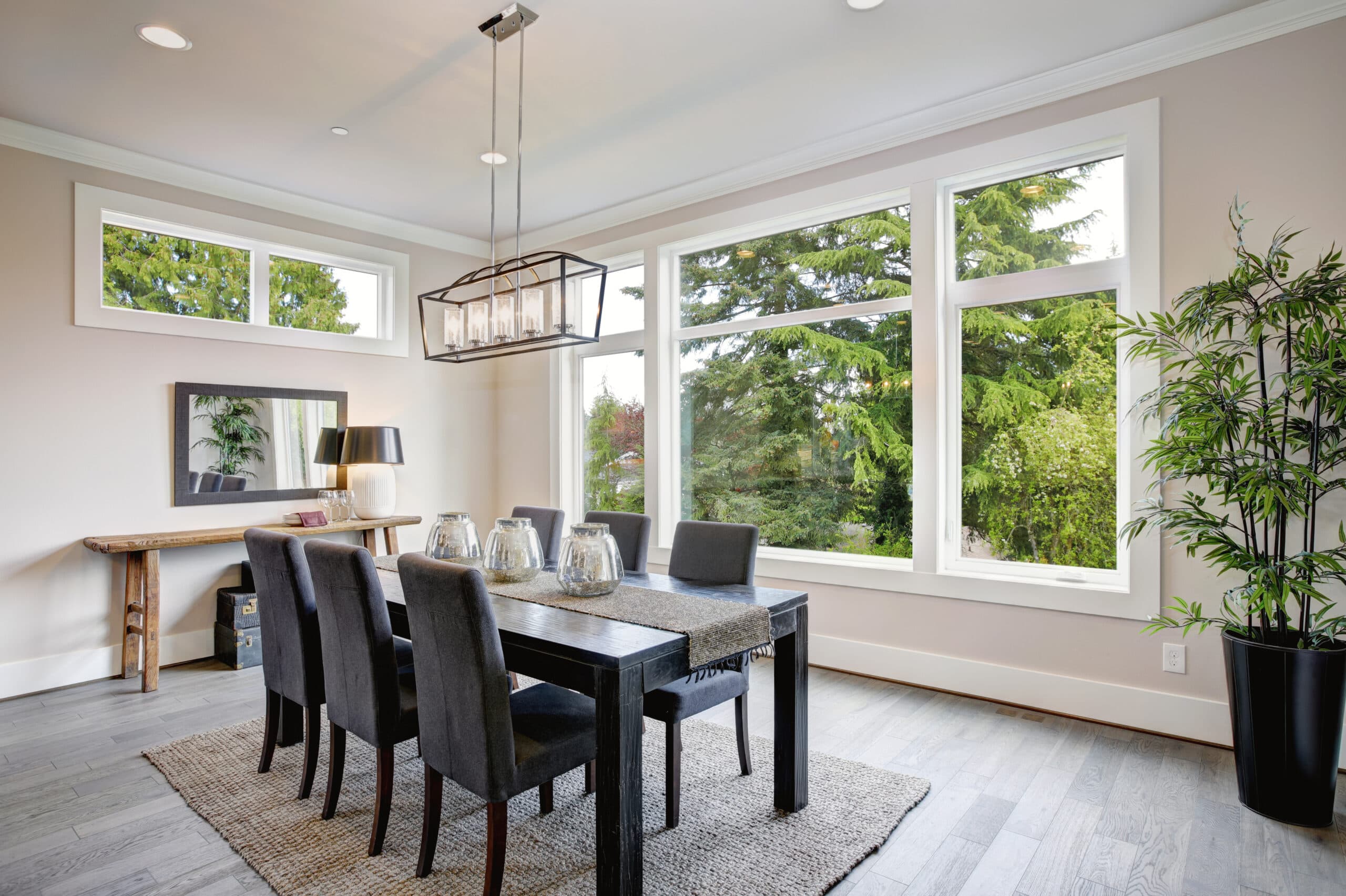 Modern dining room with large windows offering a view of lush greenery. Room features a black dining table surrounded by six gray upholstered chairs, a woven rug, and a wooden side table with a lamp and mirror. Ceiling has a rectangular chandelier.