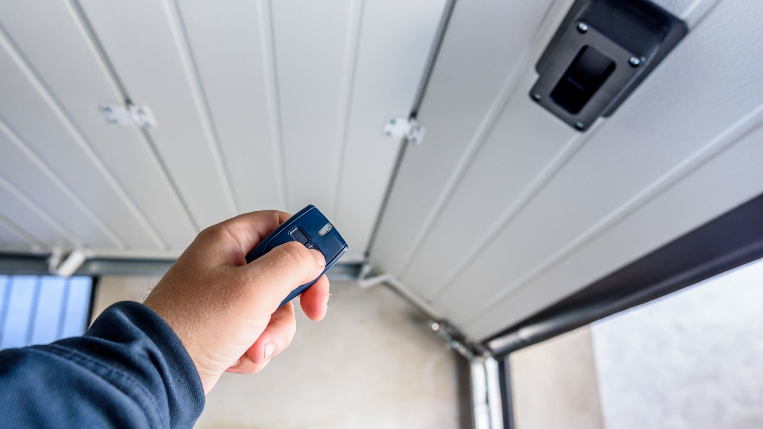 A close-up of a person's hand holding a remote control directed towards an opening garage door. The person is pressing a button on the remote. The scene is inside the garage, with the door partially open and the garage's interior visible in the background.