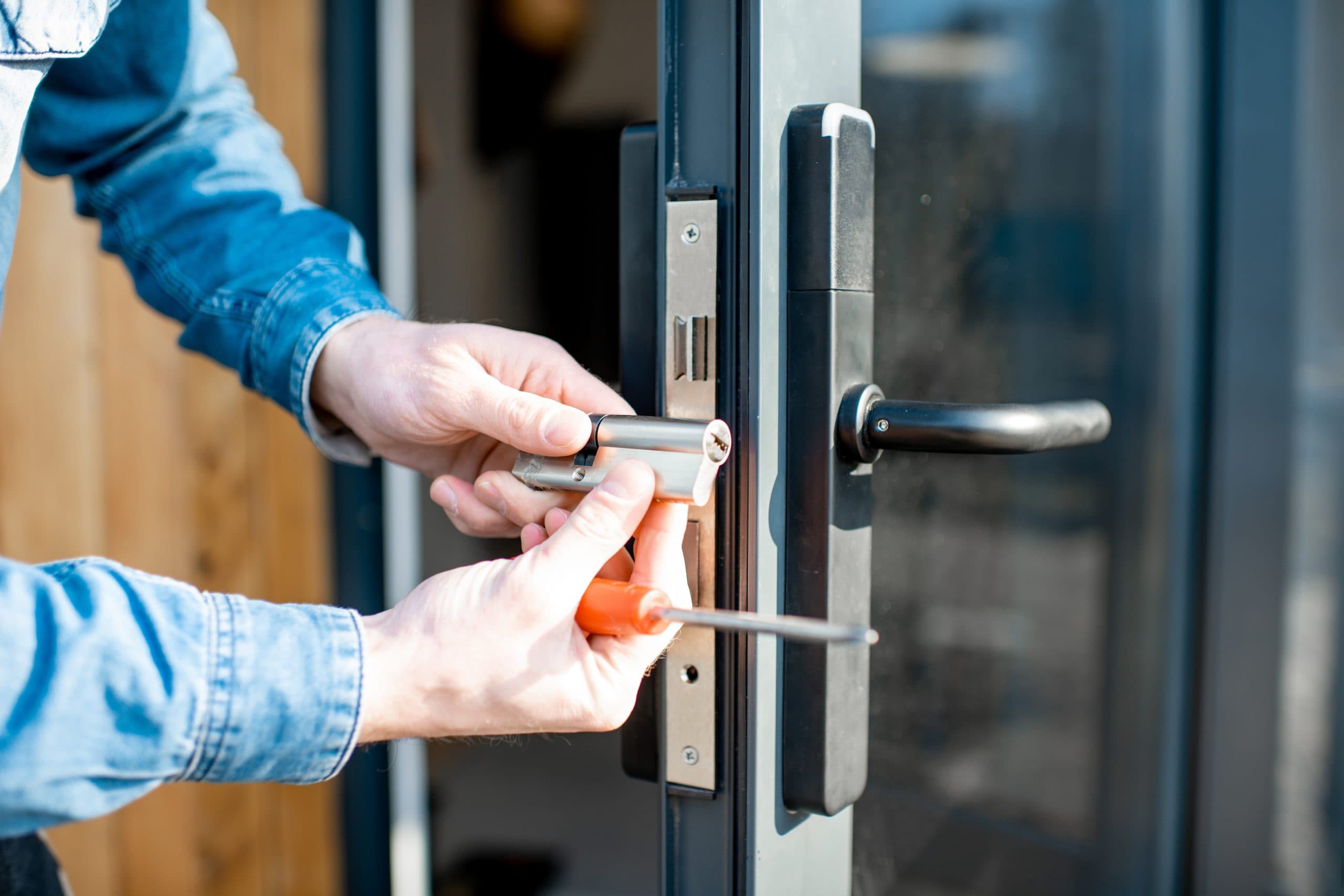 A person in a denim shirt is installing or repairing a silver door lock on a glass door. They are holding a cylindrical component of the lock in one hand and a screwdriver in the other hand. The door is black with a black handle.