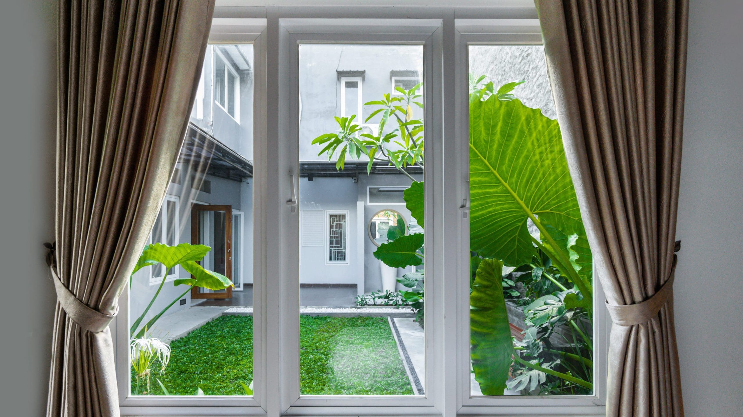 A view through a large window with beige curtains, showing a lush, green courtyard with various plants and a small lawn. The courtyard is surrounded by a modern building with white and gray walls.