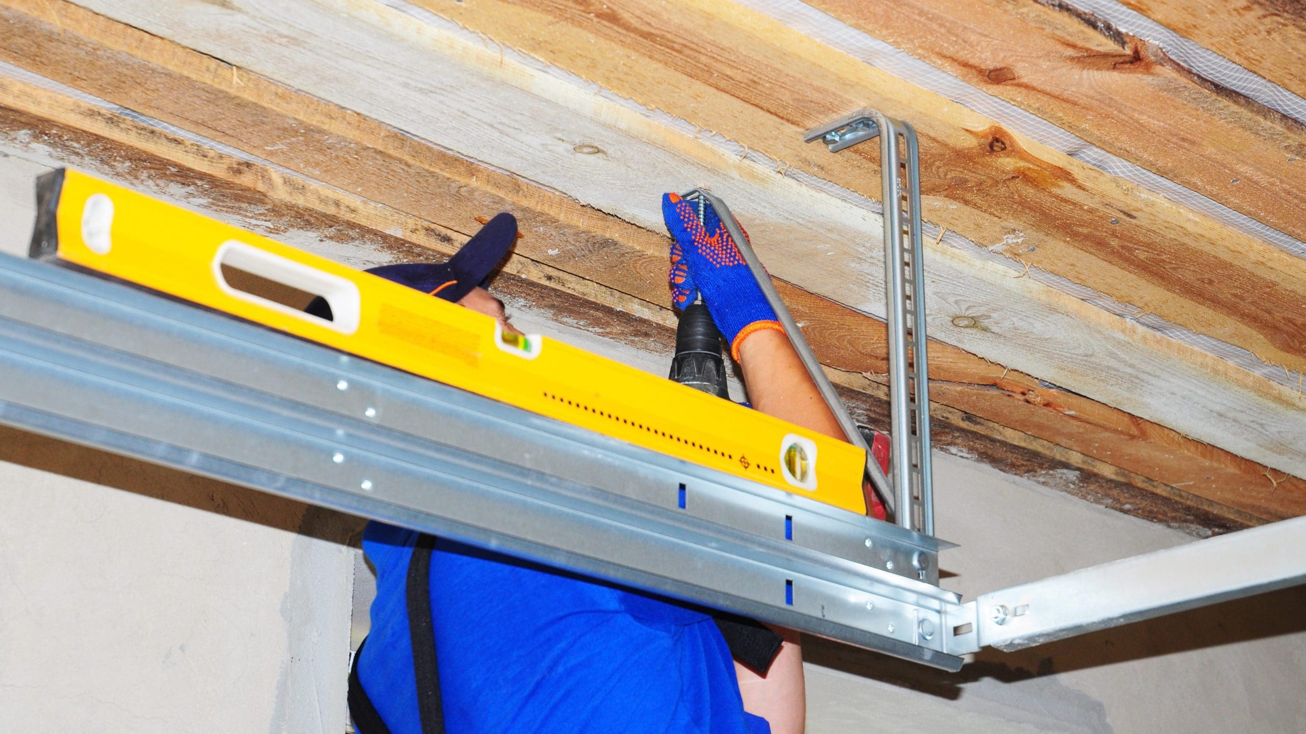 A construction worker in a blue shirt and gloves uses a power drill to attach a metal frame to a wooden ceiling. A yellow spirit level rests on the metal frame to ensure it's straight. The scene takes place indoors with a partially completed ceiling.