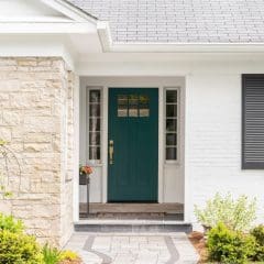 A charming house exterior featuring a front door painted in teal, flanked by glass panels and lit by sidelights. The house has white brick walls, black shutters, a stone facade on part of the structure, and a small, neatly maintained garden leading to the entrance.