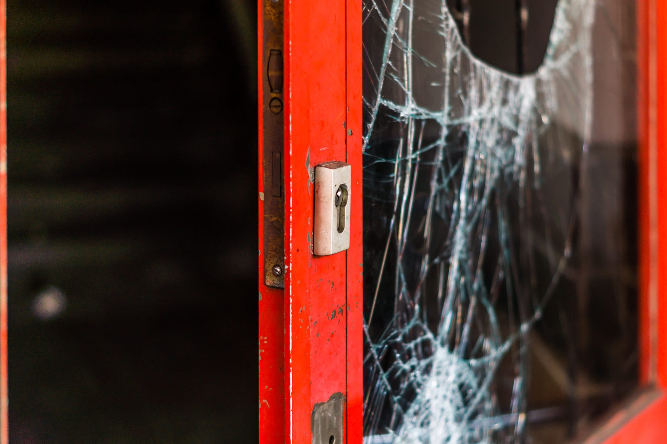 A close-up of a door painted in red with a broken glass pane. The glass has numerous cracks radiating from a hole, suggesting impact. The door’s metal lock is visible on the left. The background is dark and out of focus.