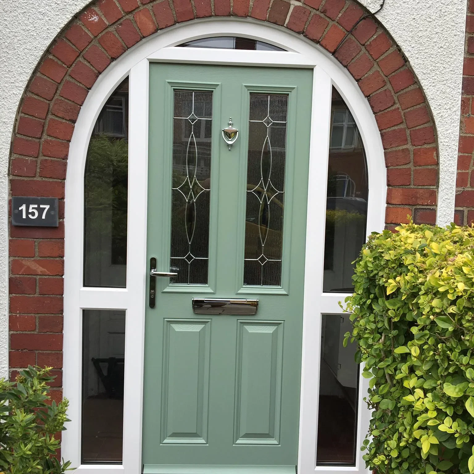 A green front door framed by a red brick archway with white trim. The door has two narrow, decorative glass panels, a mail slot, and a silver door knocker. The house number 157 is displayed on a plaque to the left. Green bushes are on the right side.