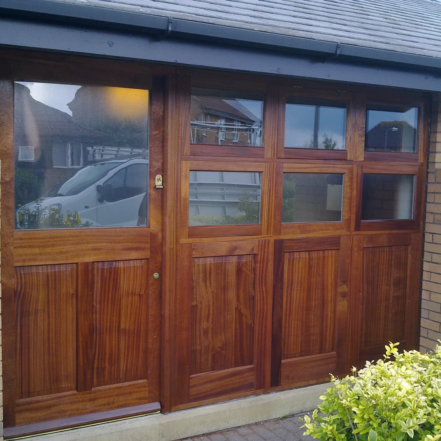 A sleek wooden garage door with multiple glass panels on the upper half is set into a brick exterior. The door reflects sunlight, highlighting its polished finish. A parked white van is slightly visible through the glass panels. Green shrubs line the area in front.